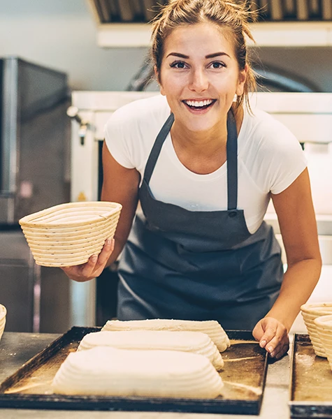 Smiling woman in apron holds basket preparing dough loaves on trays in bakery kitchen.
