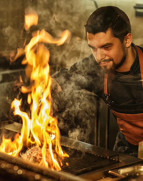 Chef grilling meat on a flaming grill with intense flames and smoke in a kitchen.
