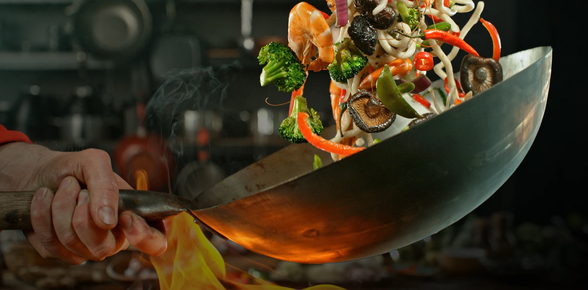 Hand tossing shrimp and vegetable stir-fry in a flaming wok in a kitchen.