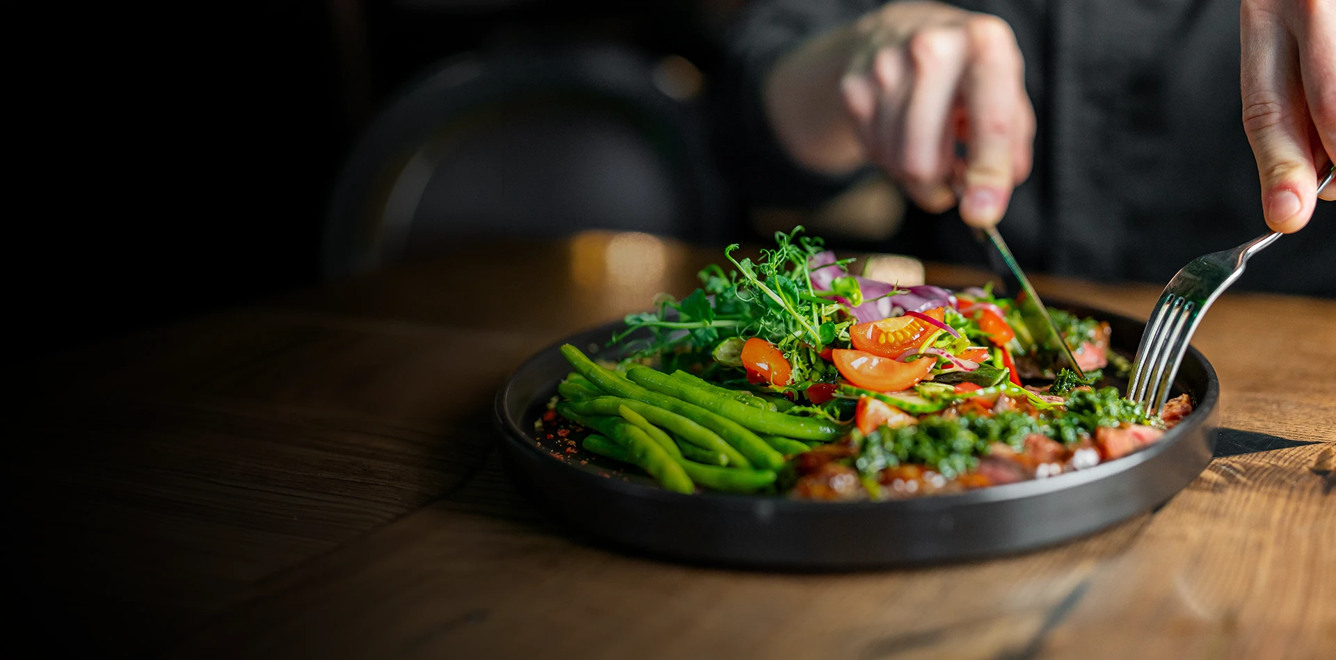 Person eating chimmichurri steak topped with green herbs, served with green beans and fresh salad on black plate.