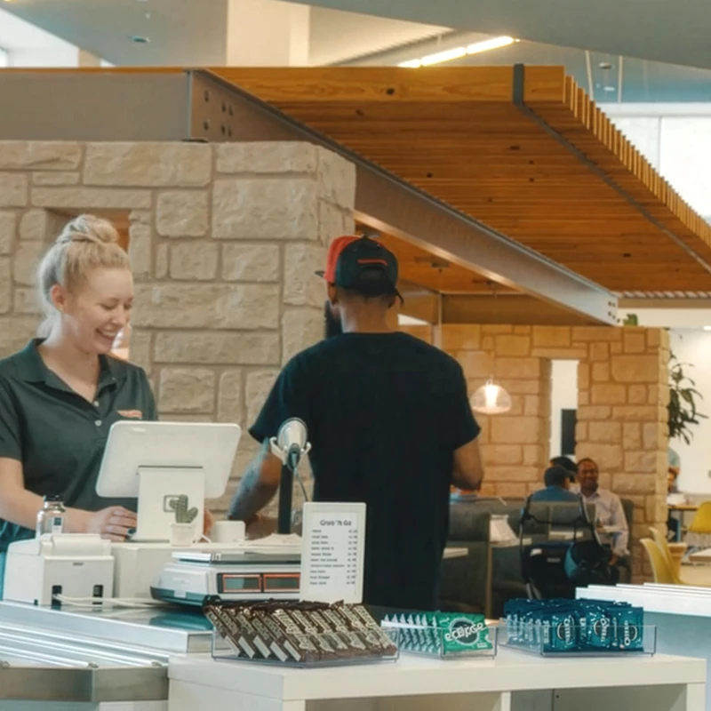 Smiling female cashier at counter serving a customer in a modern cafe setting with snacks displayed.