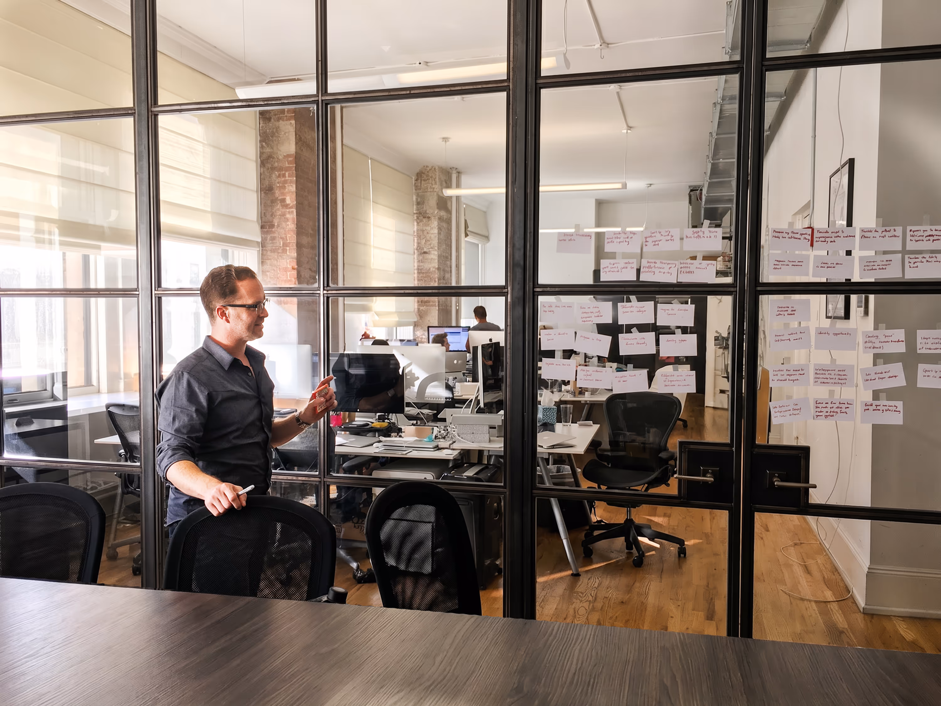 Man in glasses standing by glass wall with sticky notes in modern office with desks and computers.
