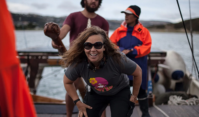 A woman leans forward directly at the camera smiling while wearing sunglasses and a headphones around her neck. Two other people stand behind her. They are all on a boat and the water is out of focus in the distance.
