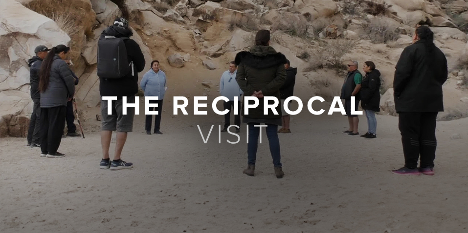 A group of people stand in a circle attentively in a sandy rocky area