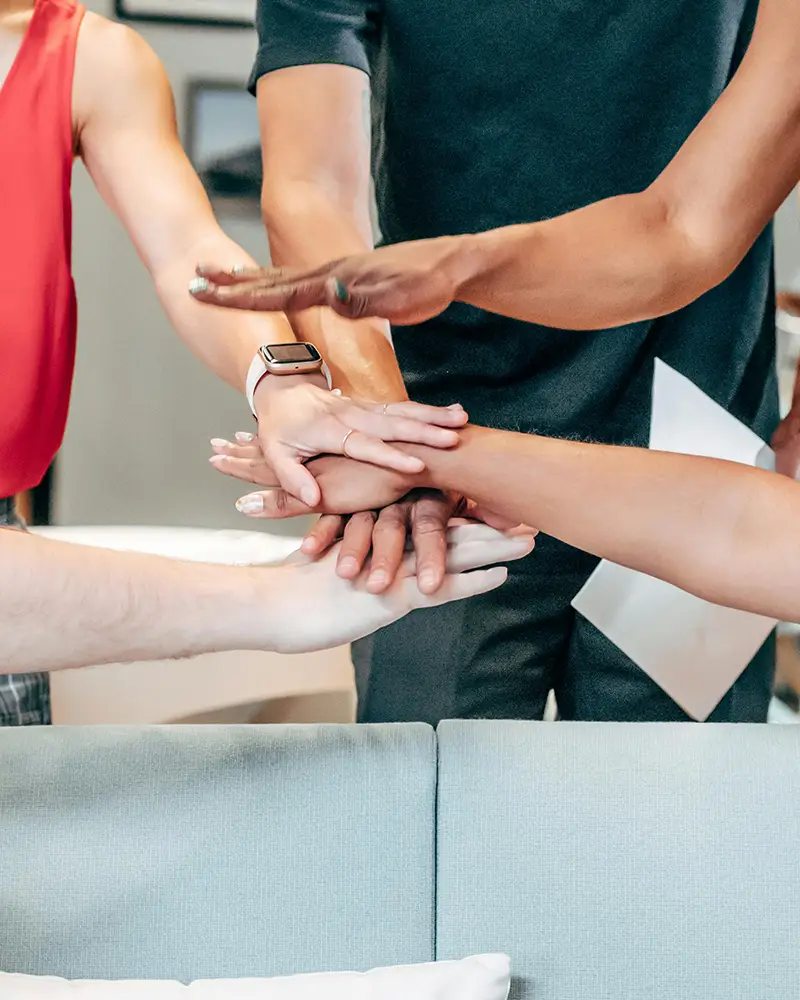 A group of people, standing in a circle, each has a hand outstretched towards the center, overlapping, like a huddle at a sports game. 