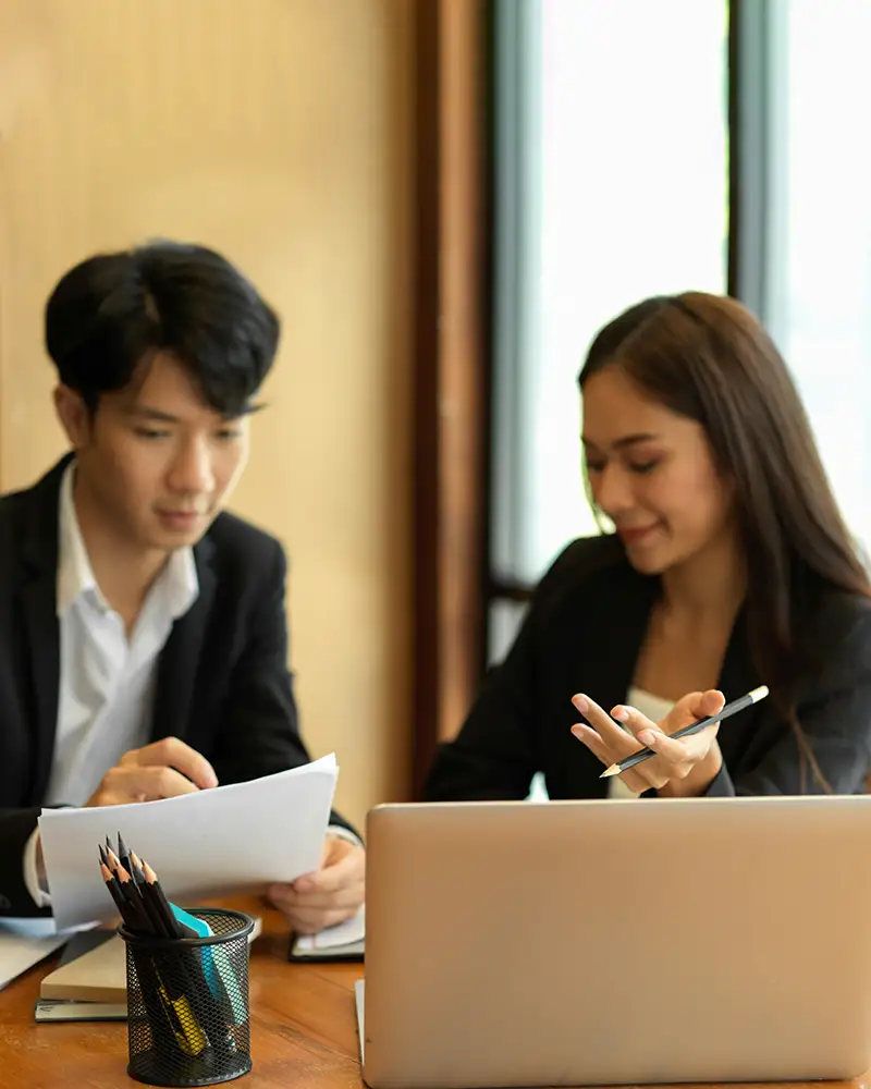Two people sit at a computer in a professional office setting. They are reviewing a document in front of them, with a laptop open nearby.