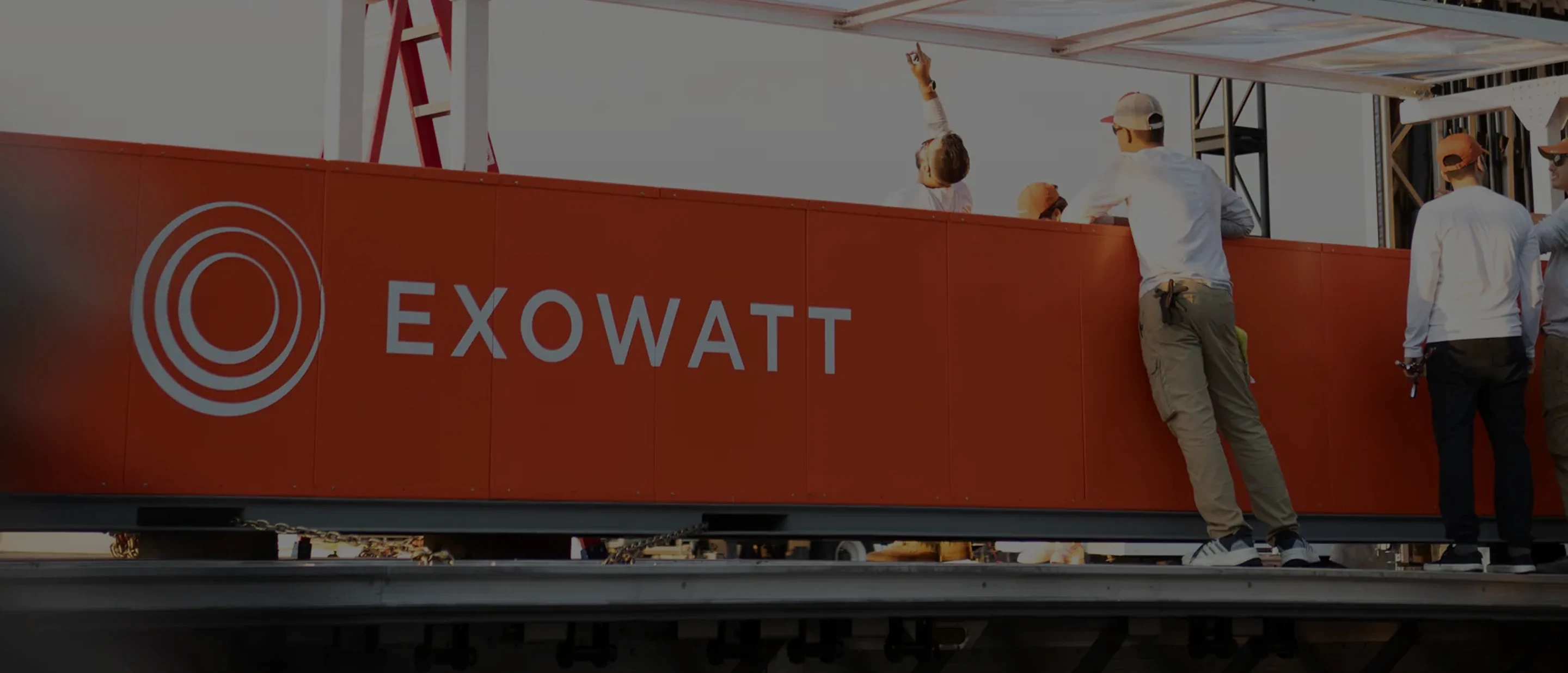 Group of workers in white shirts and caps gathered beside a large red Exowatt unit, with one person pointing upward under a canopy structure.