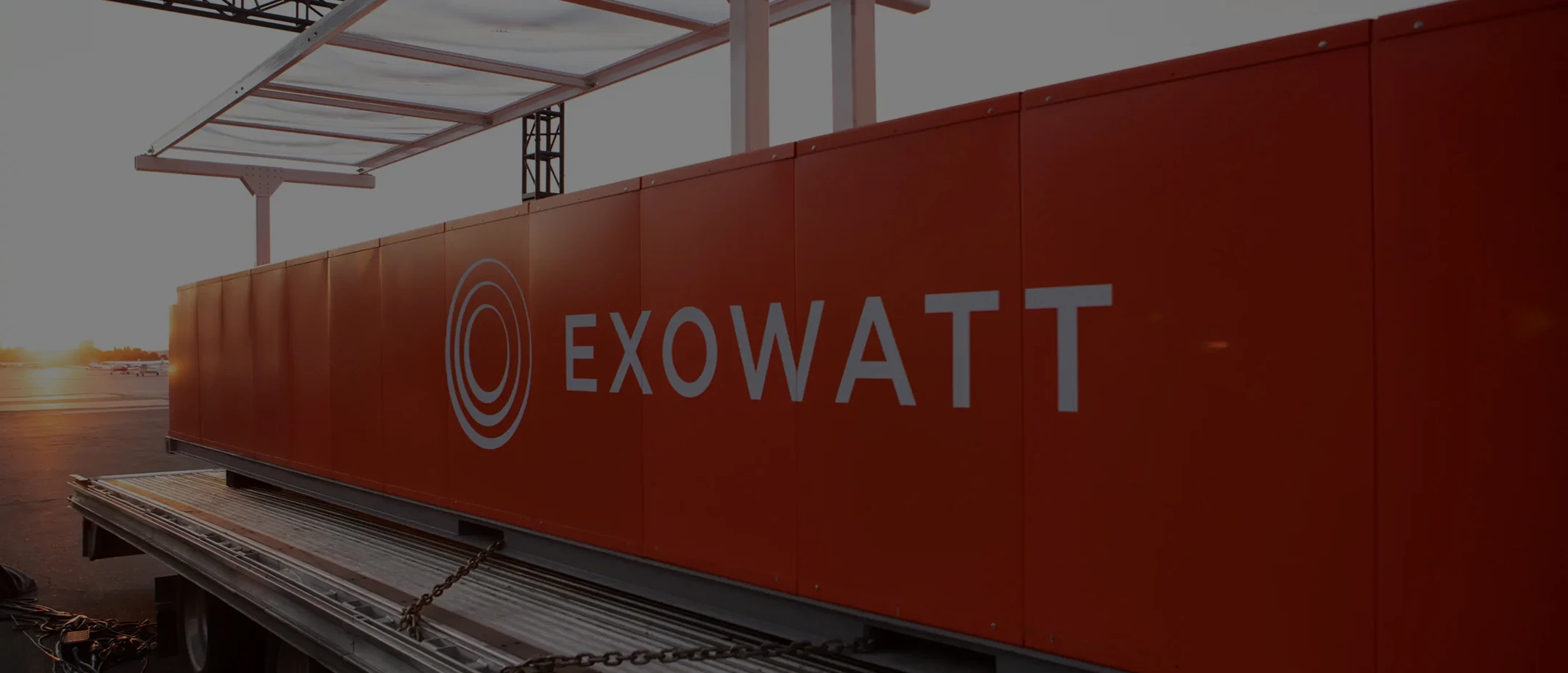 Large orange Exowatt unit mounted on a platform, with a translucent canopy frame above it and the sun setting in the background.