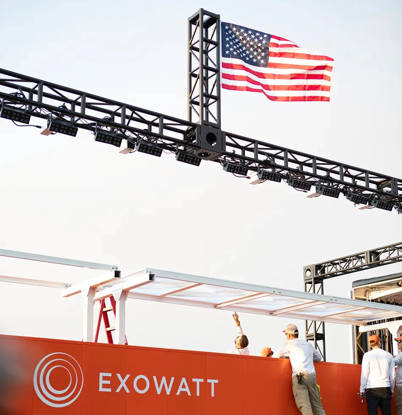 People standing beside a large red Exowatt unit while pointing upward, with an American flag waving above on a black truss structure.