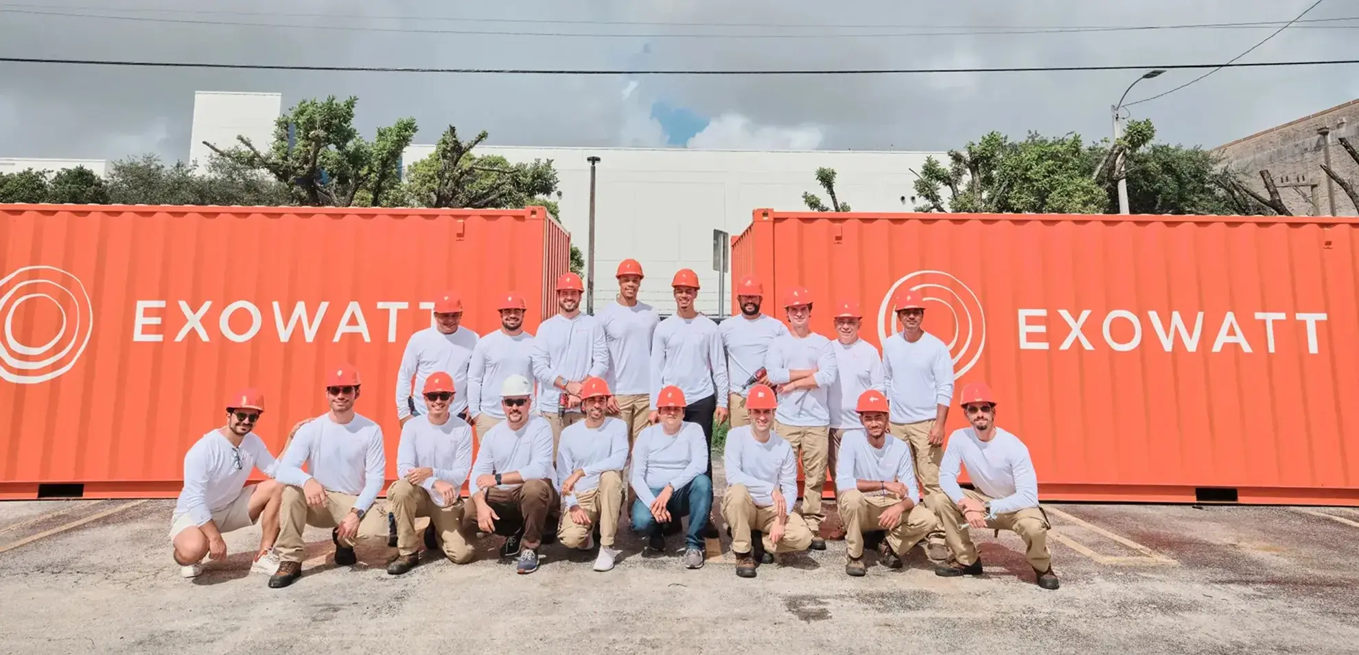 Group of Exowatt team members in white shirts and orange hard hats posing together in front of two large red Exowatt containers.