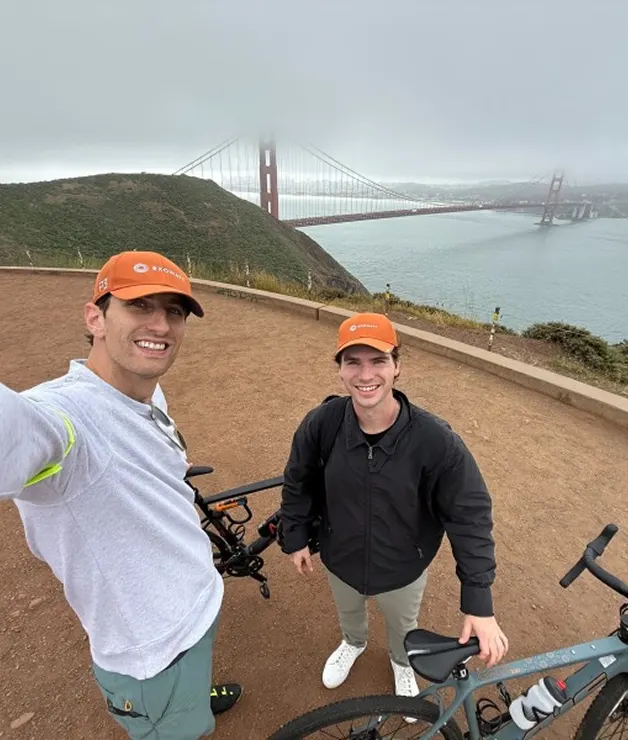 Two men wearing orange Exowatt caps posing with bicycles near the Golden Gate Bridge, partly obscured by fog.