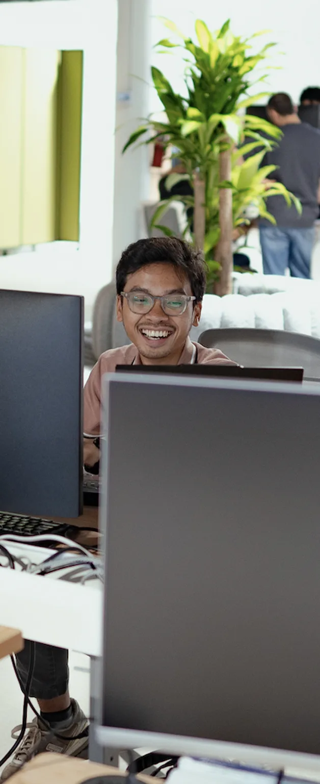 Man smiling while working at a computer desk in a bright office with multiple monitors and plants in the background.
