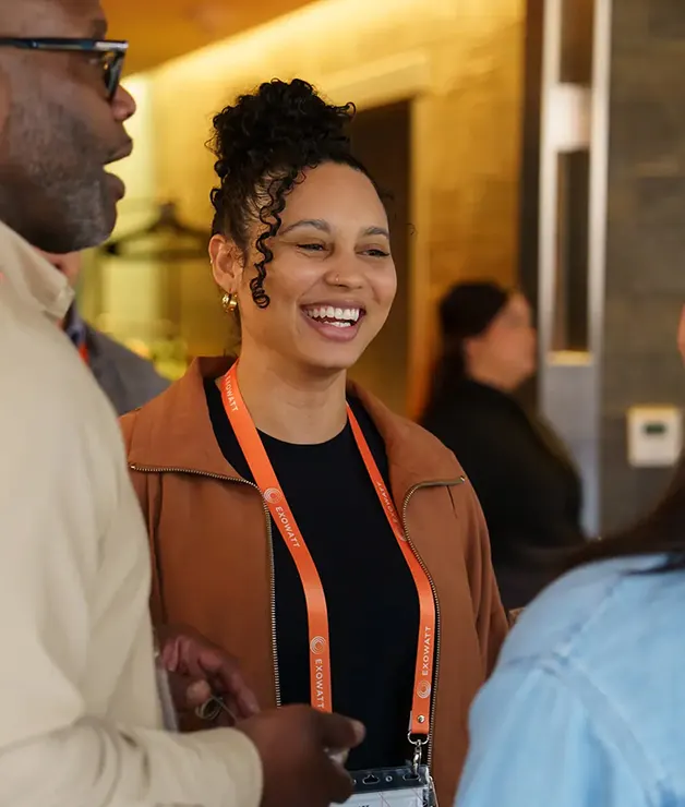 Woman smiling while wearing an orange Exowatt lanyard, chatting with others in a warmly lit indoor setting.