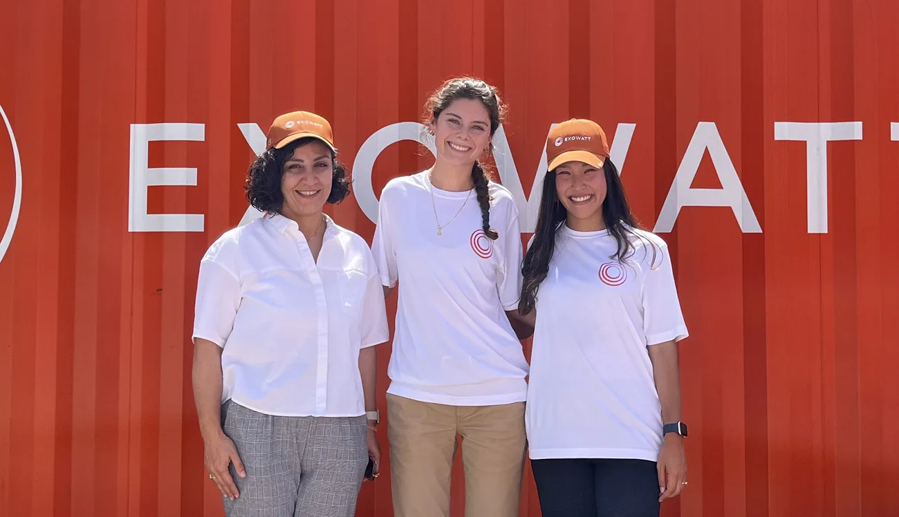 Three women smiling in front of a large red Exowatt backdrop, two wearing matching white Exowatt shirts and orange caps.