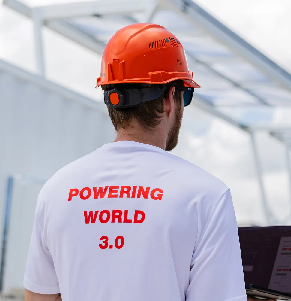 Worker in an orange hard hat and white shirt with 'Powering World 3.0' printed on the back, standing outdoors near solar equipment.