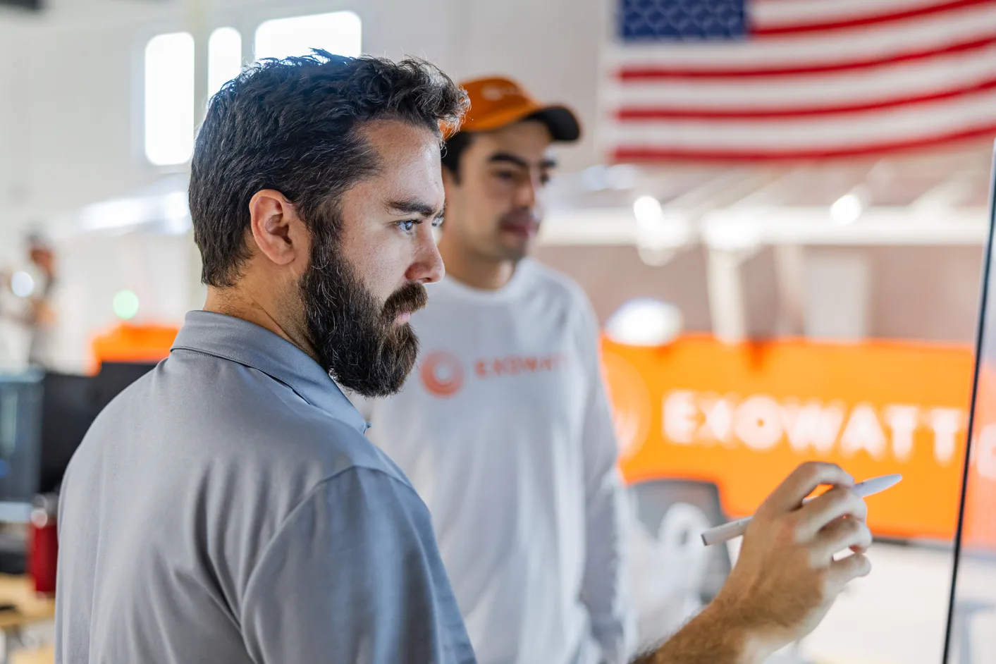 Man holding a stylus and working on a screen with focus, while another in Exowatt gear stands behind him, with an American flag and orange Exowatt equipment visible in the background.