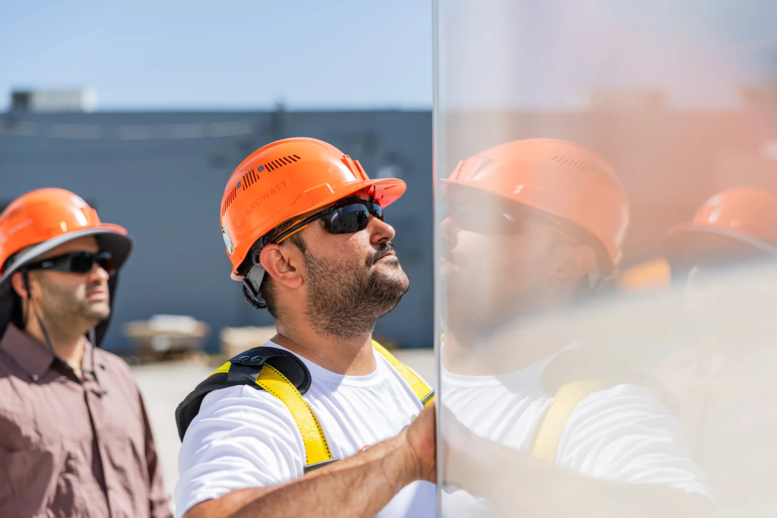 Man in an orange Exowatt hard hat and safety glasses carefully handling a reflective solar panel, with colleagues in similar gear working alongside him outdoors.