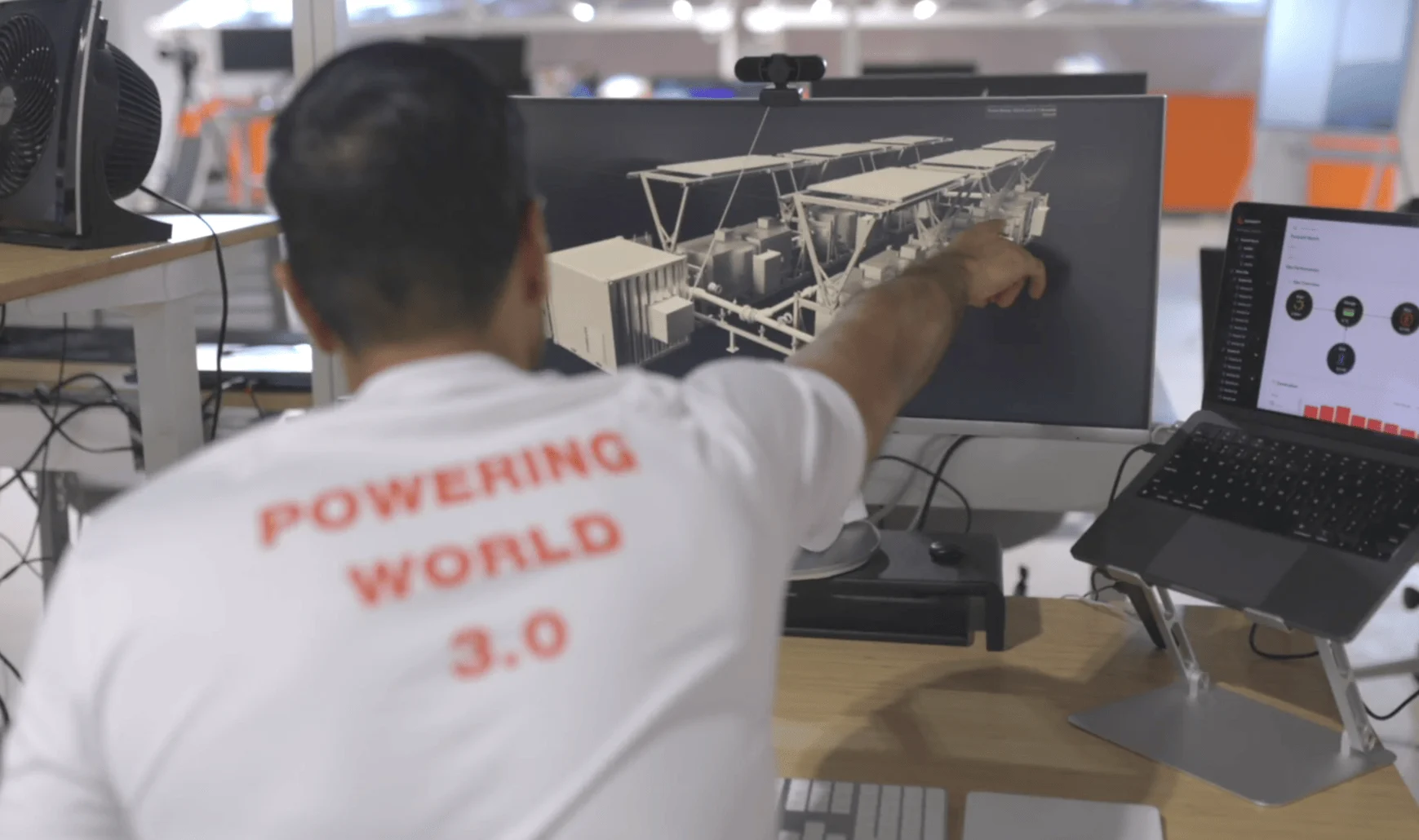 Person in a 'Powering World 3.0' shirt pointing at a computer screen displaying a 3D model of Exowatt equipment, with data charts visible on a nearby laptop.