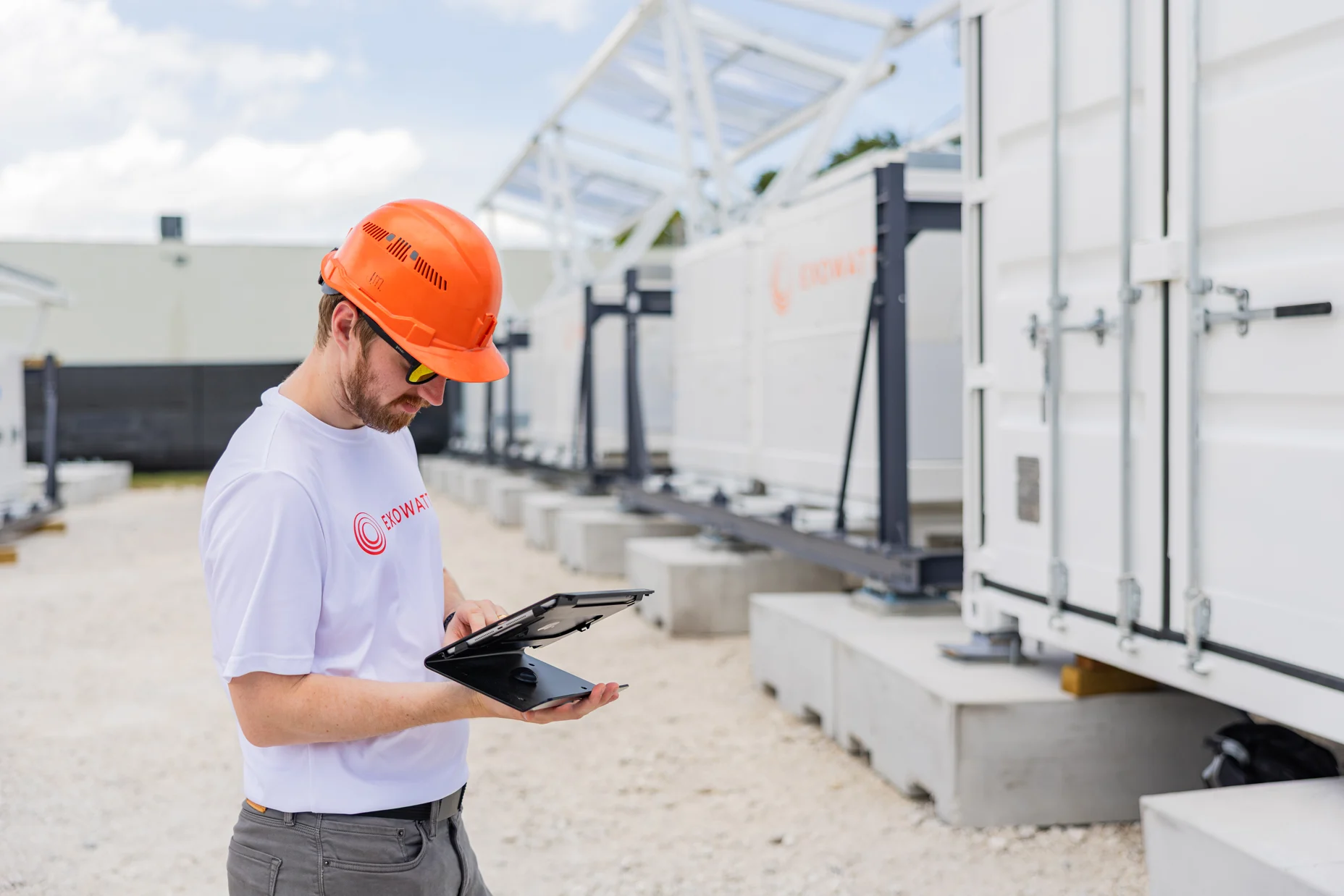 Worker in an orange hard hat and Exowatt shirt using a laptop outdoors, standing beside rows of white modular Exowatt units on concrete platforms.
