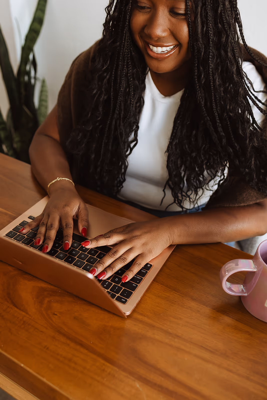Smiling woman with braided hair typing on a rose gold laptop at a wooden table with a pink mug nearby.