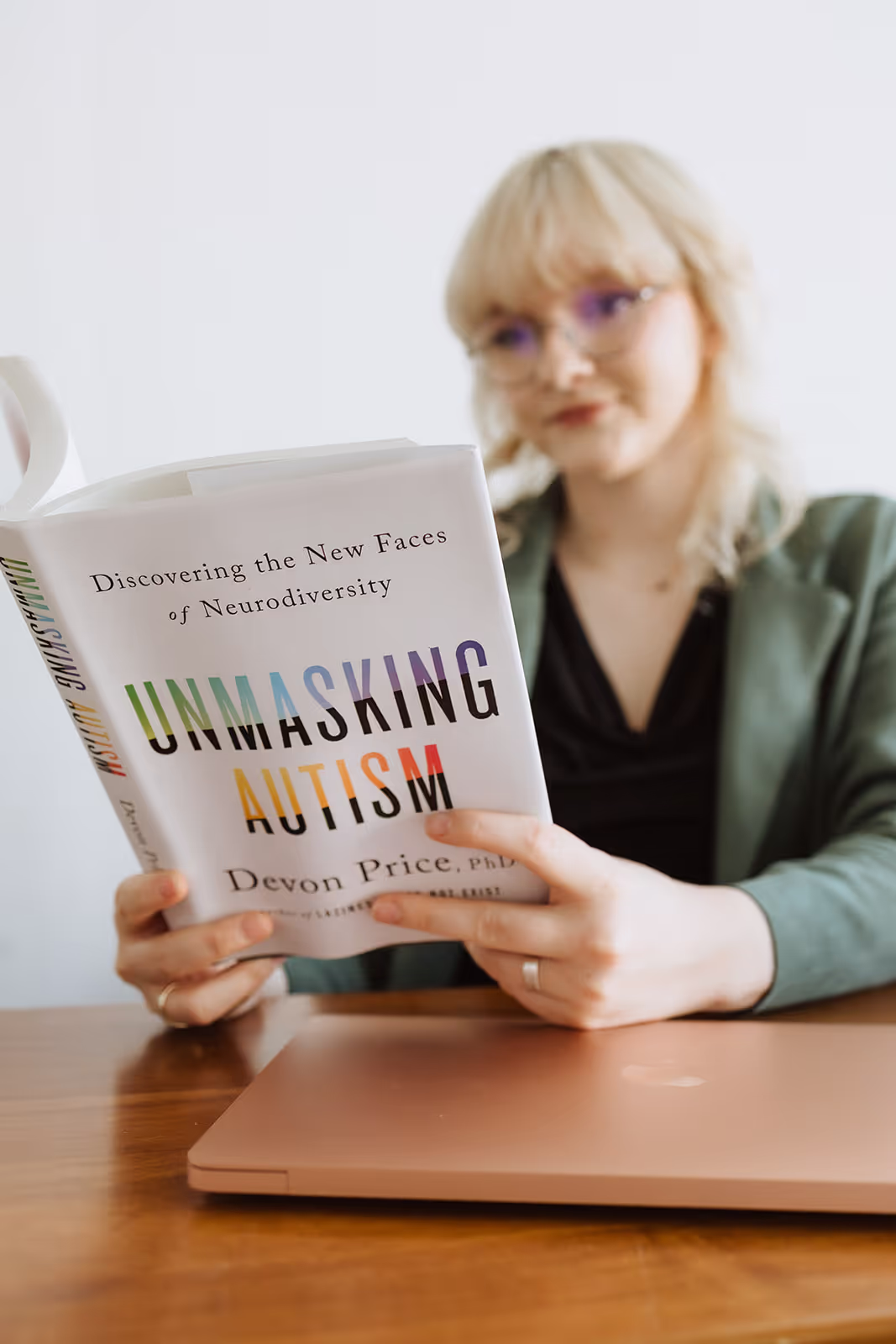 Person with blonde hair and glasses reading the book 'Unmasking Autism' by Devon Price at a wooden table with a closed laptop.