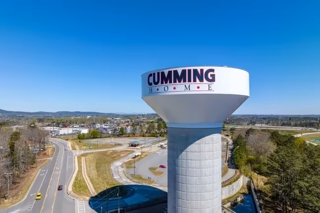White water tower with "Cumming" text overlooking suburban road and landscape.