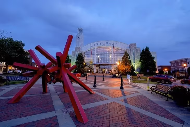 Large red sculpture in front of city hall at twilight with brick plaza.