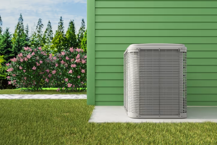 Air conditioning unit outside green house with flowering bush and trees.