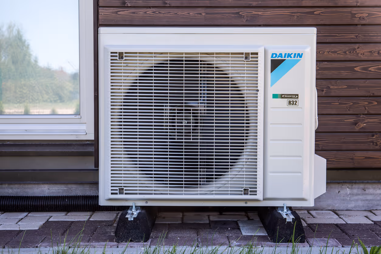 A white Daikin heat pump unit with a black fan and blue and black logo is installed on concrete blocks on a patio, positioned against a dark wood-paneled house next to a white window.