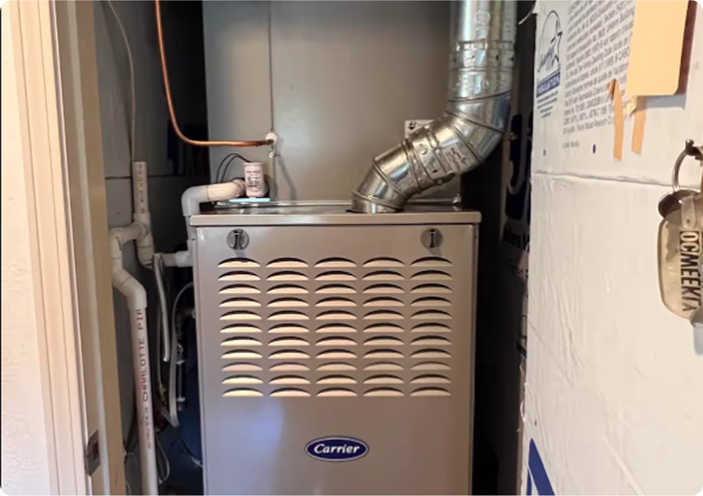 A close-up of a gray Carrier furnace with a white label, connected to silver ducts and copper pipes, is positioned in a narrow utility space.