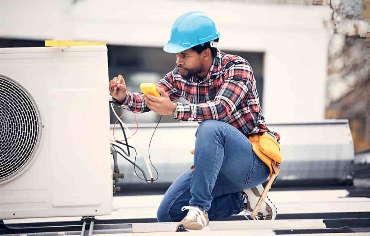 Technician in blue helmet checking electrical connections near air conditioning unit