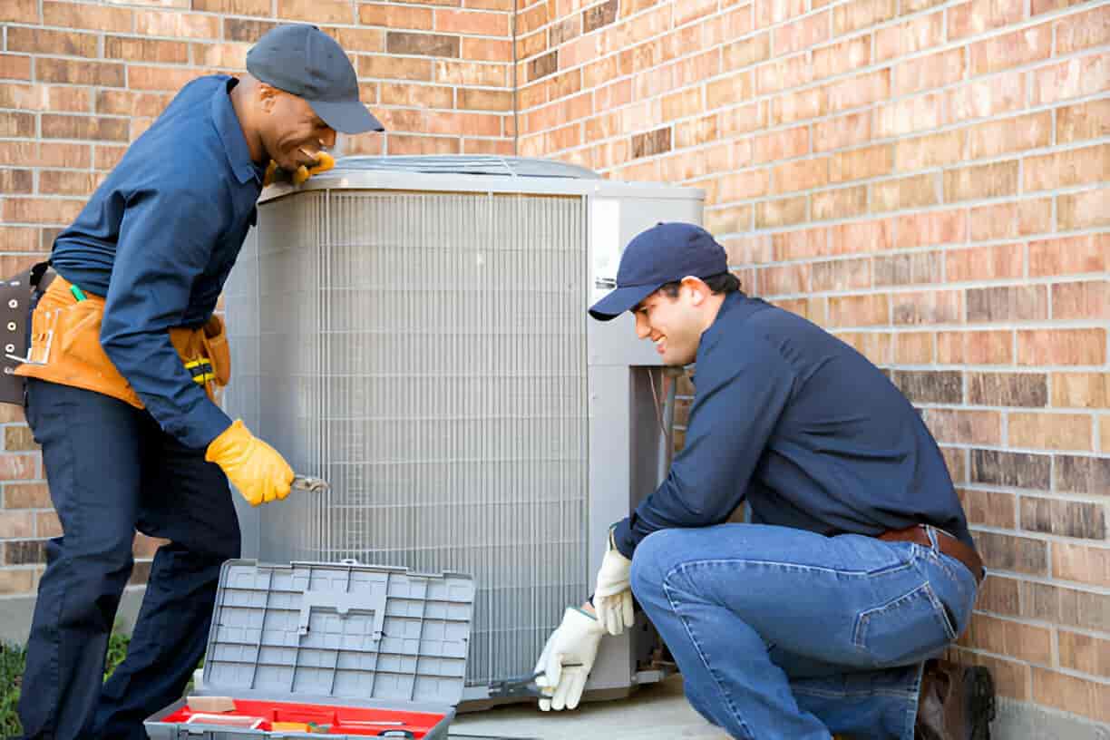 Two technicians repairing an air conditioning unit outside a brick building