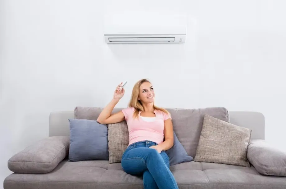  A woman on a sofa pointing a remote at a wall-mounted AC unit.