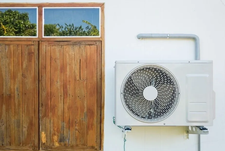 An outdoor AC unit mounted on a white wall next to a wooden window.