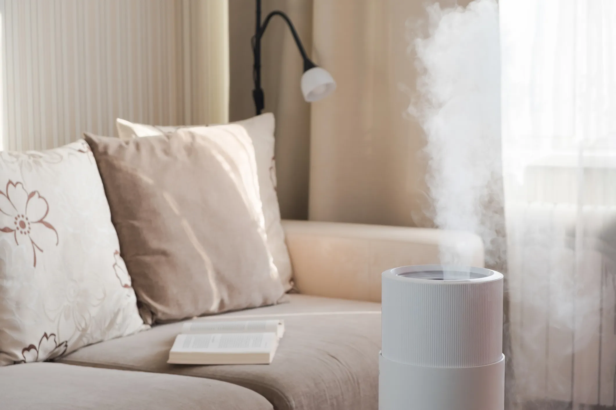 Air purifier emitting mist near cozy sofa with book and decorative pillows