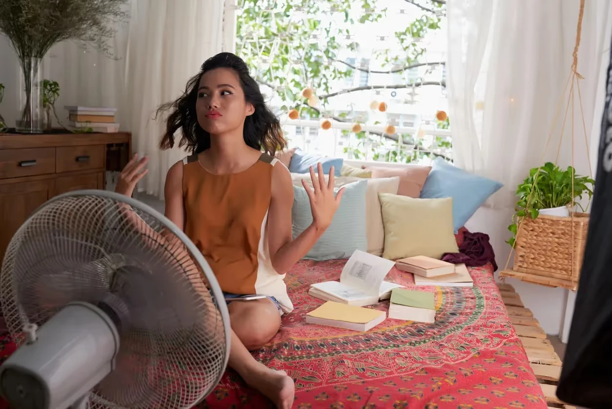 Woman sitting on floor with books, using fan to cool off
