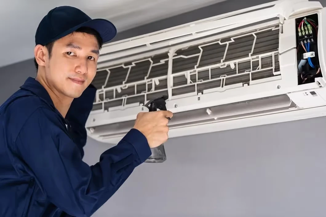 Technician in blue uniform cleaning air conditioning unit with spray