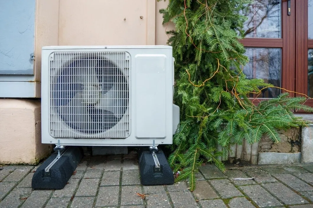 An outdoor heat pump or AC unit on risers next to a building and evergreen plant.