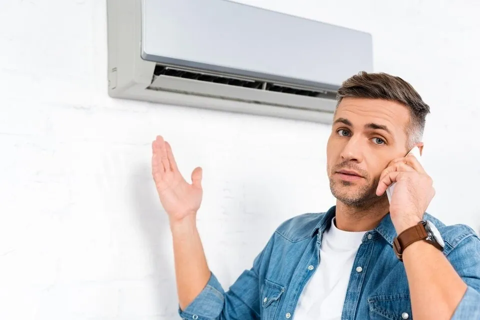 A man on a phone call, gesturing in frustration under an AC unit.