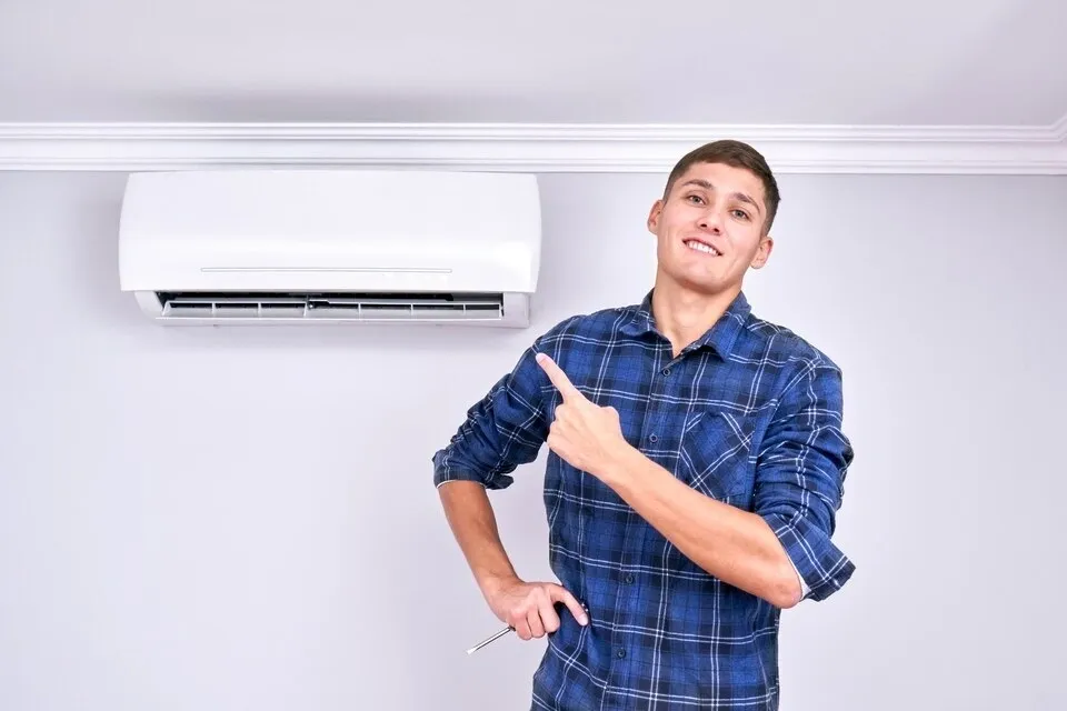 A man in a blue plaid shirt points to a wall-mounted AC unit.