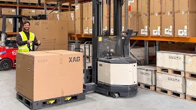 Worker in a yellow vest loads a large XAG branded box onto a warehouse forklift.