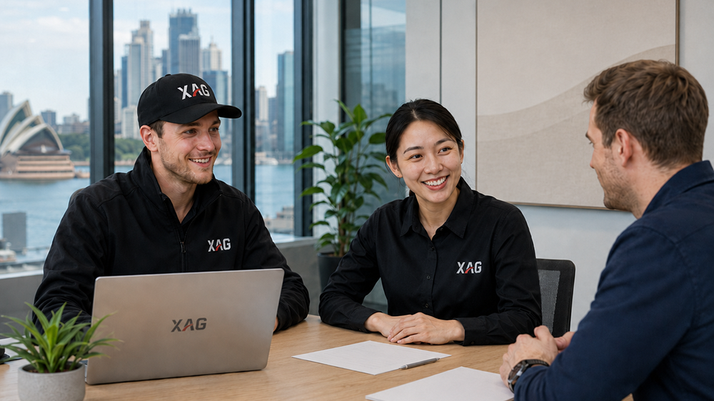 Three people smiling and talking at a table with laptops and papers in an office with city view.