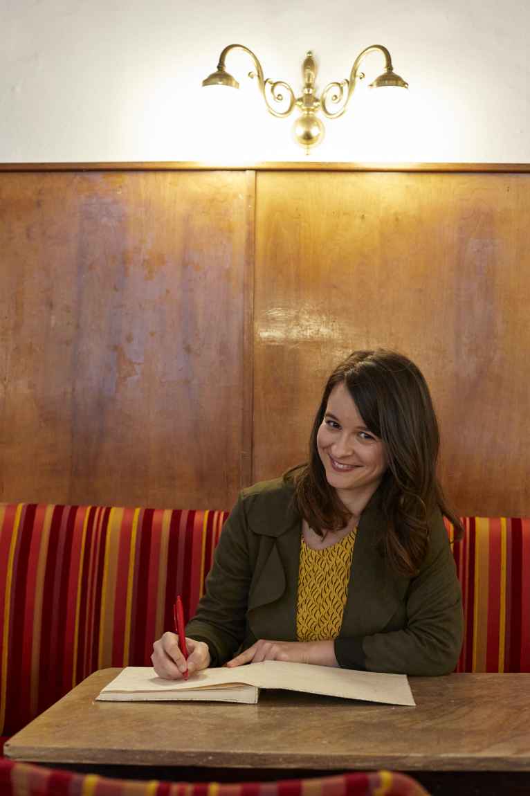 Marlene Groihofer von audioportait sitzt an einem Tisch und schreibt etwas in ihr Notizbuch. Dabei lächelt sie sympathisch in die Kamera.
