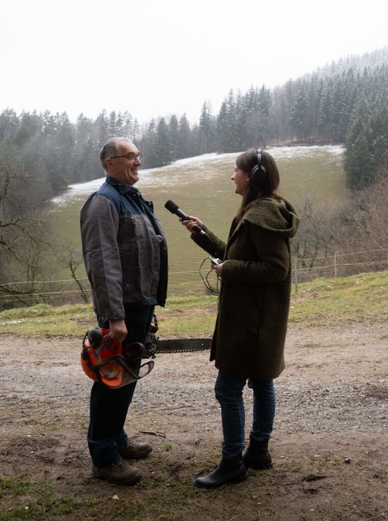 Marlene Groihofer von audioportait interviewt einen Holzarbeiter mit Motersäge im Freien. Im Hintergrund sieht man schöne Landschaft mit Wiesen, Hügeln und schneebedeckte Bäume. 