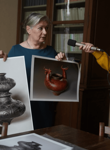 Eine ältere Dame spricht in das Mikrofon von audioportait und zeigt dabei verschiedene Bilder mit Tonvasen und Skulpturen.