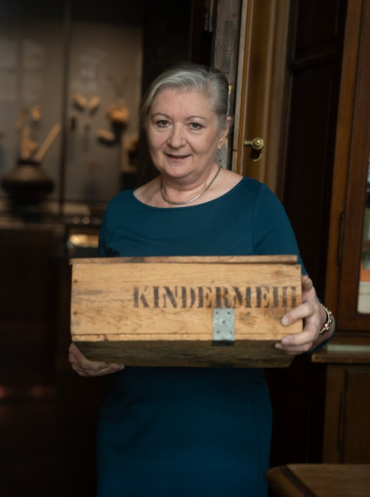 Ein Bilder einer älteren Dame welche eine Holzkiste in ihren Händen hält. Sie trägt ein Blaues Stoffkleid und sieht sehr zufrieden aus.