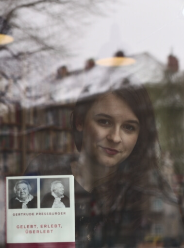Marlene Groihofer von audioportrait sieht mit einem Buch in ihrer Hand durch nach außen durch ein Fenster. Man sieht Reflexionen von Gebäuden in dem Fenster. 