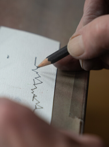Eine Nahaufnahme von einer Hand welche mit einem Bleistift etwas auf einen Zettel schreibt.