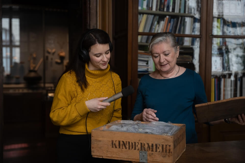 Marlene Groihofer von audioportrait interviewt eine Dame in einem wohnlichen Raum mit einer Holzkiste im Vordergrund und Holzschränke mit vielen Büchern im Hintergurnd.