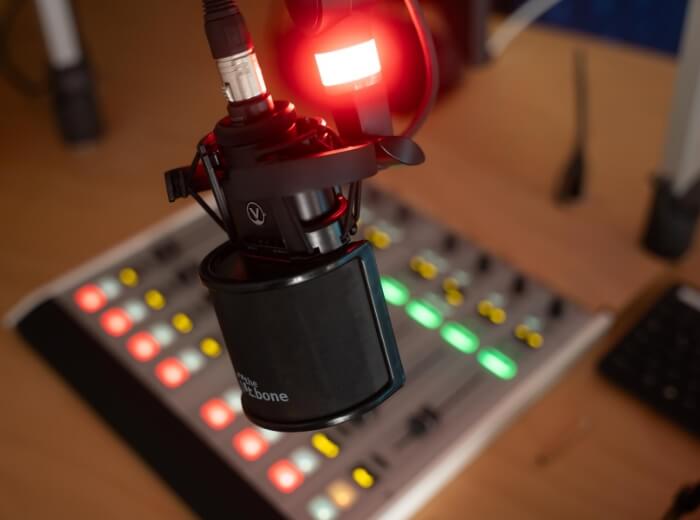 Eine Ansicht von einem Mikrofon welches am Stativ über einem Mischpult hängt. Ein rotes Licht leuchtet am Mikro, welches signalisiert, dass nun Audio aufgenommen wird.